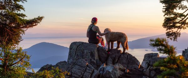 Woman and her dog sitting on a large rock over looking mountains at sunset